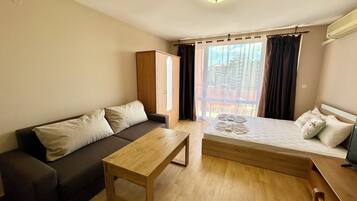 Una camera moderna con un divano, un tavolo in legno, un letto con asciugamani e vista sugli edifici attraverso ampie finestre.