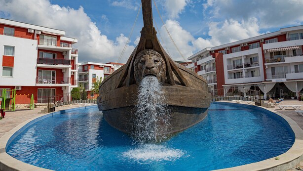 A fountain with a lion's head in the center of a swimming pool, surrounded by multi-story buildings.