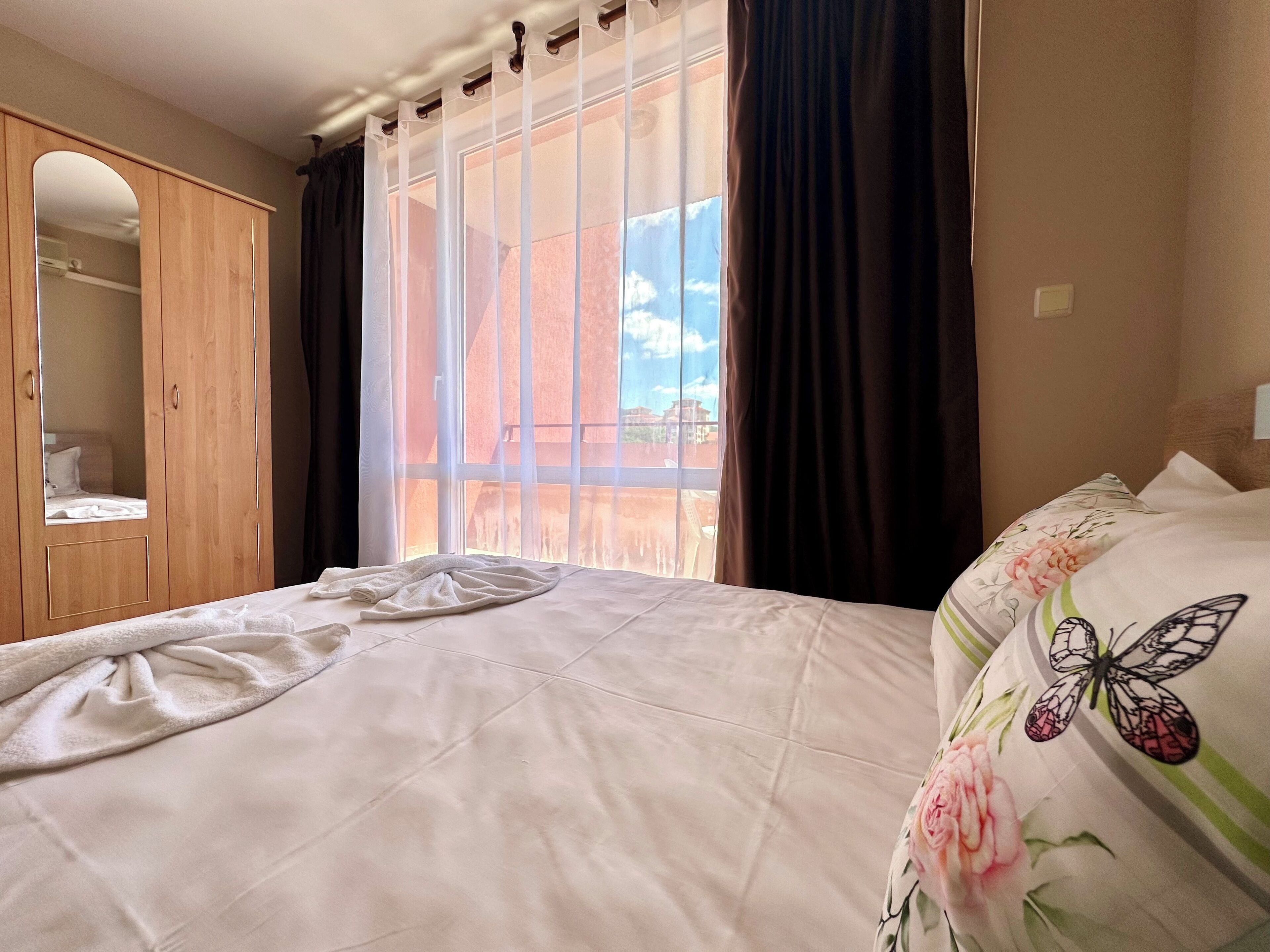 A bedroom with a large window, a wooden wardrobe, and a bed with floral bedding.
