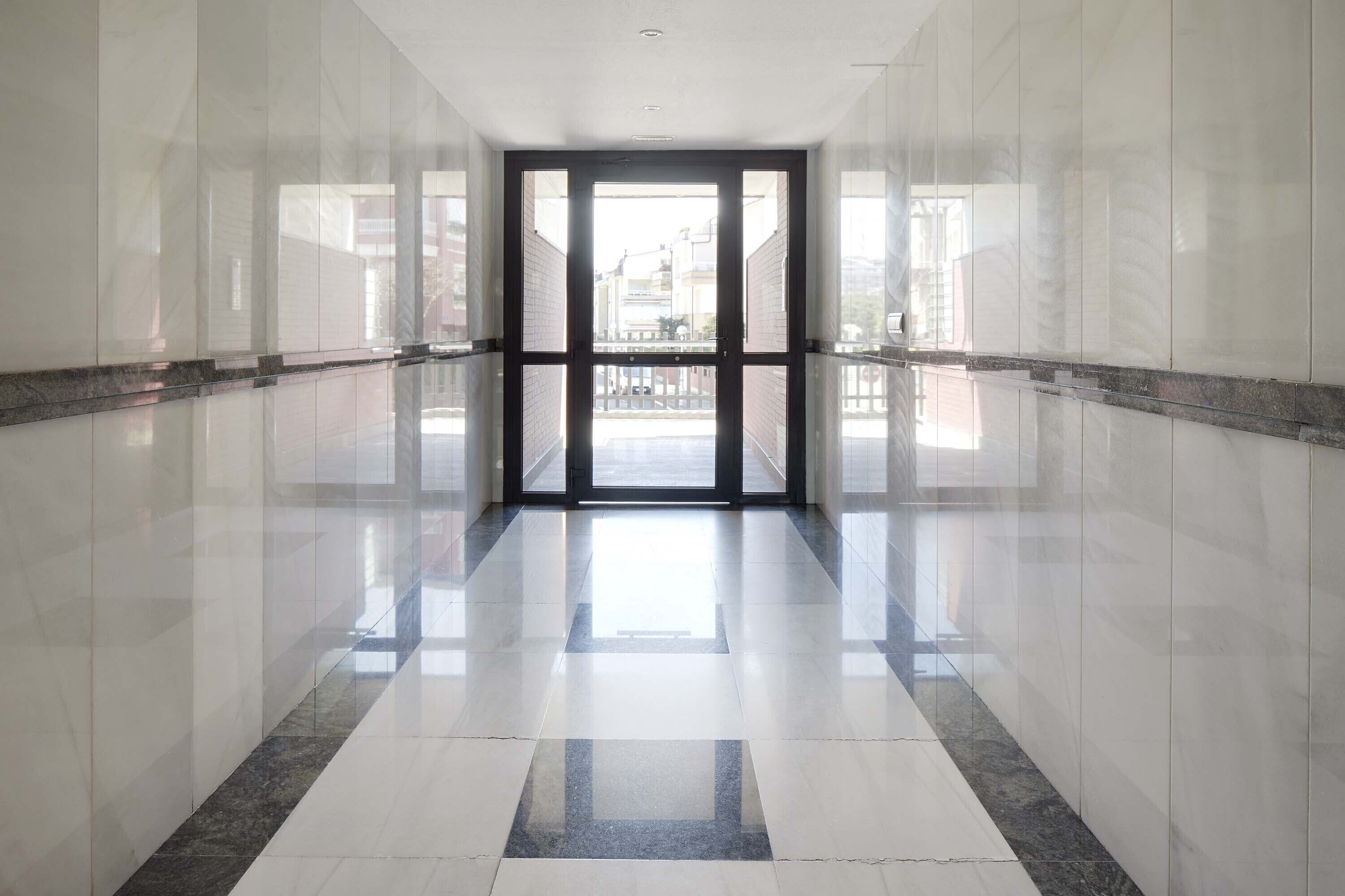 A corridor with reflective tiled floors and walls, leading to a glass door with a view of the outside.