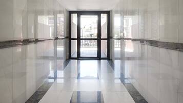A corridor with reflective tiled floors and walls, leading to a glass door with a view of the outside.