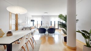 Un salon moderne avec un coin repas, comprenant une grande table blanche, deux fauteuils sombres et une plante en pot.