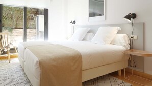 Une chambre moderne avec un grand lit, deux tables de chevet, une lampe fixée au mur et une vue sur l’extérieur, accessible par une porte vitrée.
