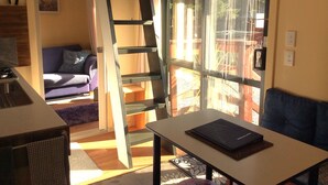Une pièce comprenant un canapé, une table avec un ordinateur portable, une échelle et une porte-fenêtre coulissante donnant sur un espace extérieur.