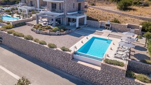 Una casa moderna de dos pisos con piscina, mobiliario exterior y un muro de piedra alrededor de la propiedad.