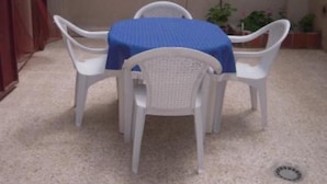 Un pequeño patio al aire libre con una mesa blanca y cuatro sillas blancas, cubierto por un mantel azul.