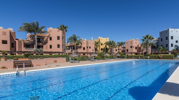 Una piscina con sillones de descanso y palmeras, rodeada de edificios de arquitectura tradicional.
