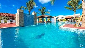 A clear blue pool with lounge chairs, palm trees, and a covered area. - Casa Alessandra 2bdr (Oranjestad-West)