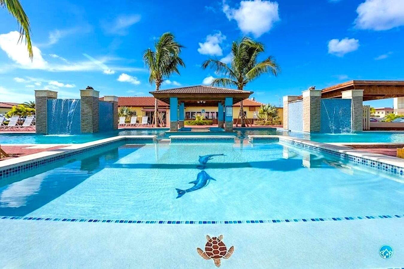 A swimming pool with a dolphin sculpture, palm trees, and a building with a tiled roof in the background.