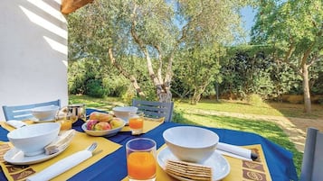 Une table de jardin, dressée pour un repas en plein air, avec des bols, des assiettes, des couverts et un verre de jus d’orange, le tout entouré de verdure et d’arbres.