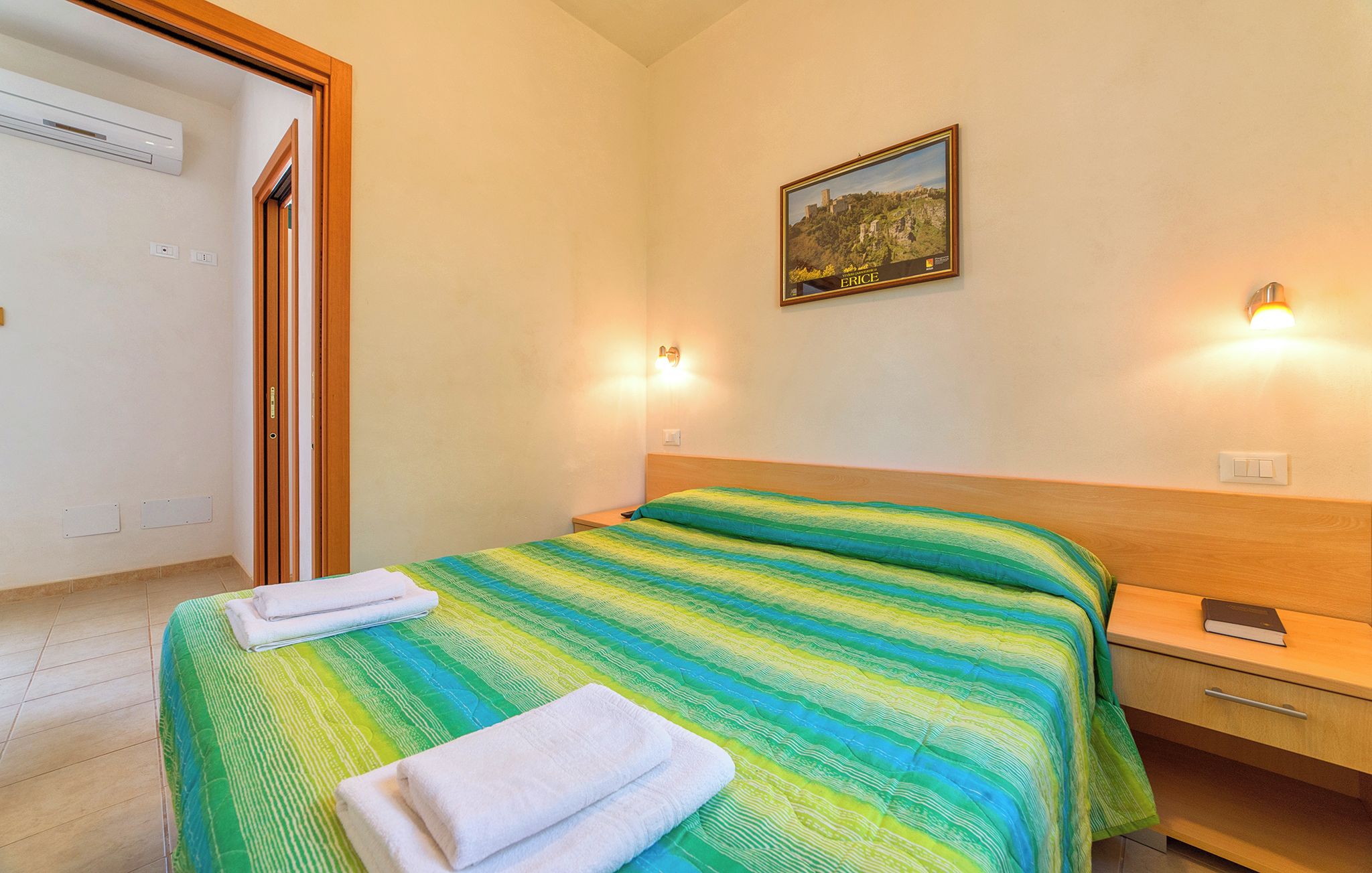 Une chambre d’hôtel avec un lit, deux lampes fixées au mur, un tableau et une table de chevet en bois.