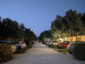 A gravel parking area with cars parked on both sides, surrounded by trees and a building with a yellow facade. - Mini cottages surrounded by nature (Marsala)