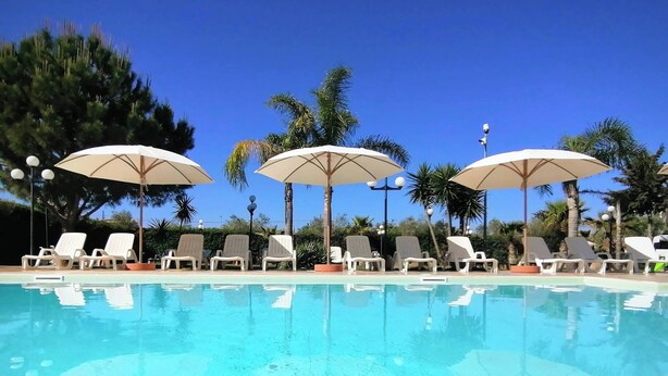 Un área de piscina con sillones de descanso y sombrillas, rodeada de palmeras.