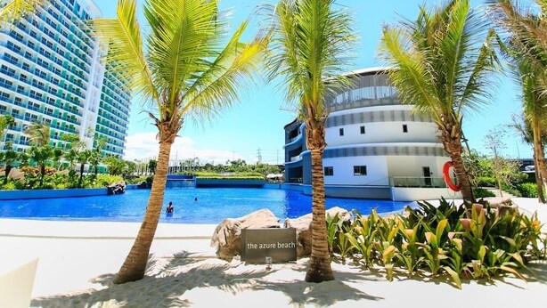 A resort pool area with palm trees, a sign reading "the azure beach," and modern buildings in the background. - Azure Tropical Paradise in your Backyard (Parañaque)