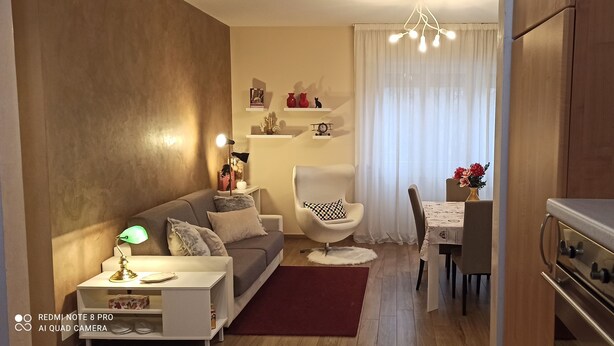 Uma sala de estar moderna com um sofá, uma mesa lateral com abajur, uma área de jantar com mesa e cadeiras, e uma cozinha com fogão.