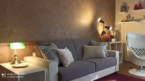 Uma sala de estar moderna com um sofá cinzento, uma poltrona branca e uma pequena mesa lateral com uma luminária. Há prateleiras com objetos decorativos e um relógio na parede.