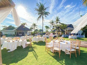 Un espacio para eventos al aire libre con mesas y sillas redondas, palmeras y luces colgantes.