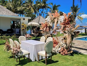 Un área de estar al aire libre con una mesa y sillas blancas, rodeada de plantas tropicales y un arco decorativo.