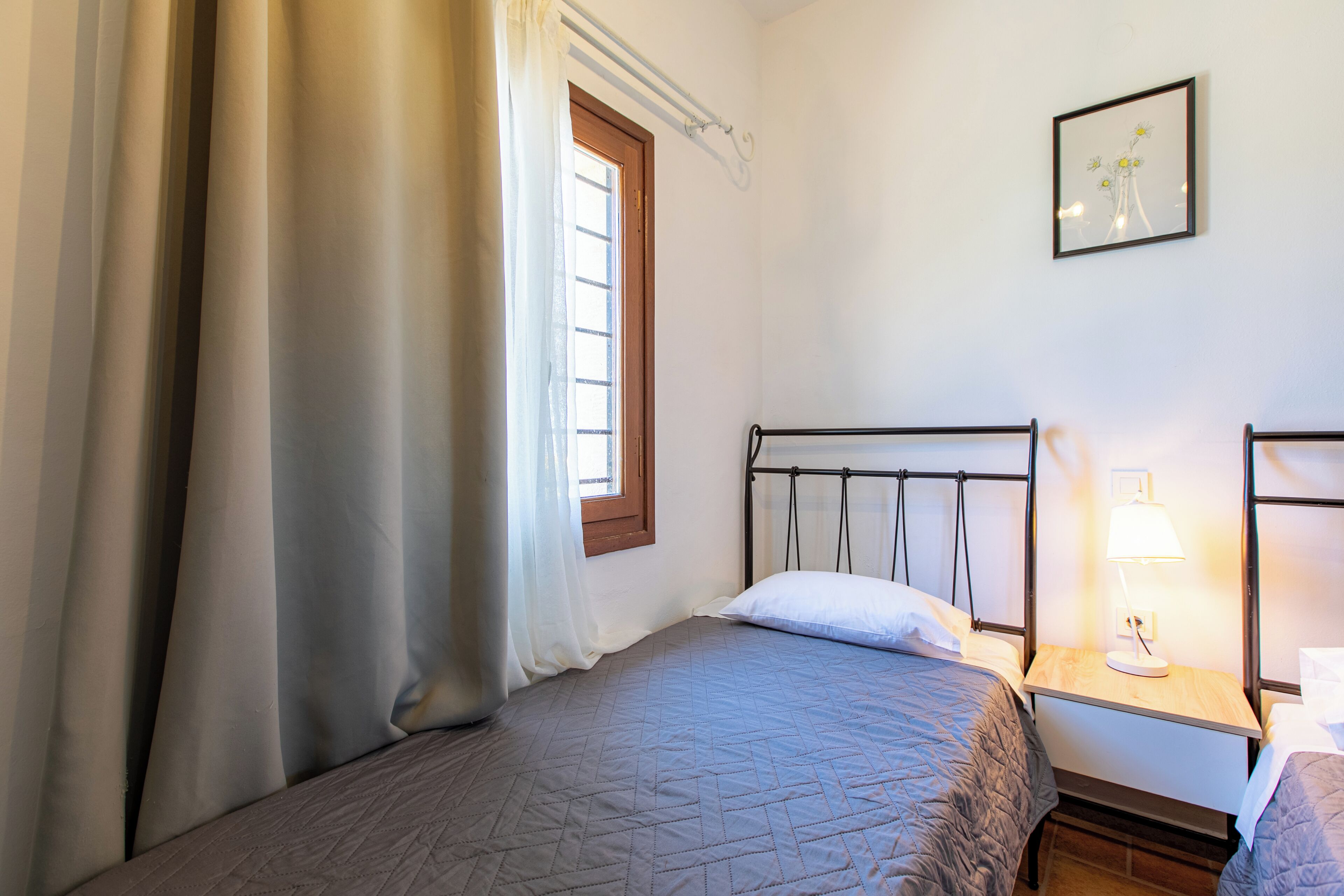 A bedroom with a single bed, a nightstand with a lamp, a window with curtains, and a framed picture on the wall.