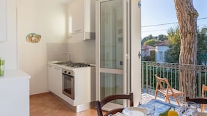 Een compacte keuken met een gasfornuis, oven en een eethoek met een tafel voor twee. Een glazen deur leidt naar een buitenbalkon met een boom en een hekwerk.