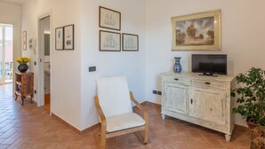 Een woonkamer met een witte fauteuil, een televisie op een wit kastje, en kunstwerken in lijsten aan de muur.