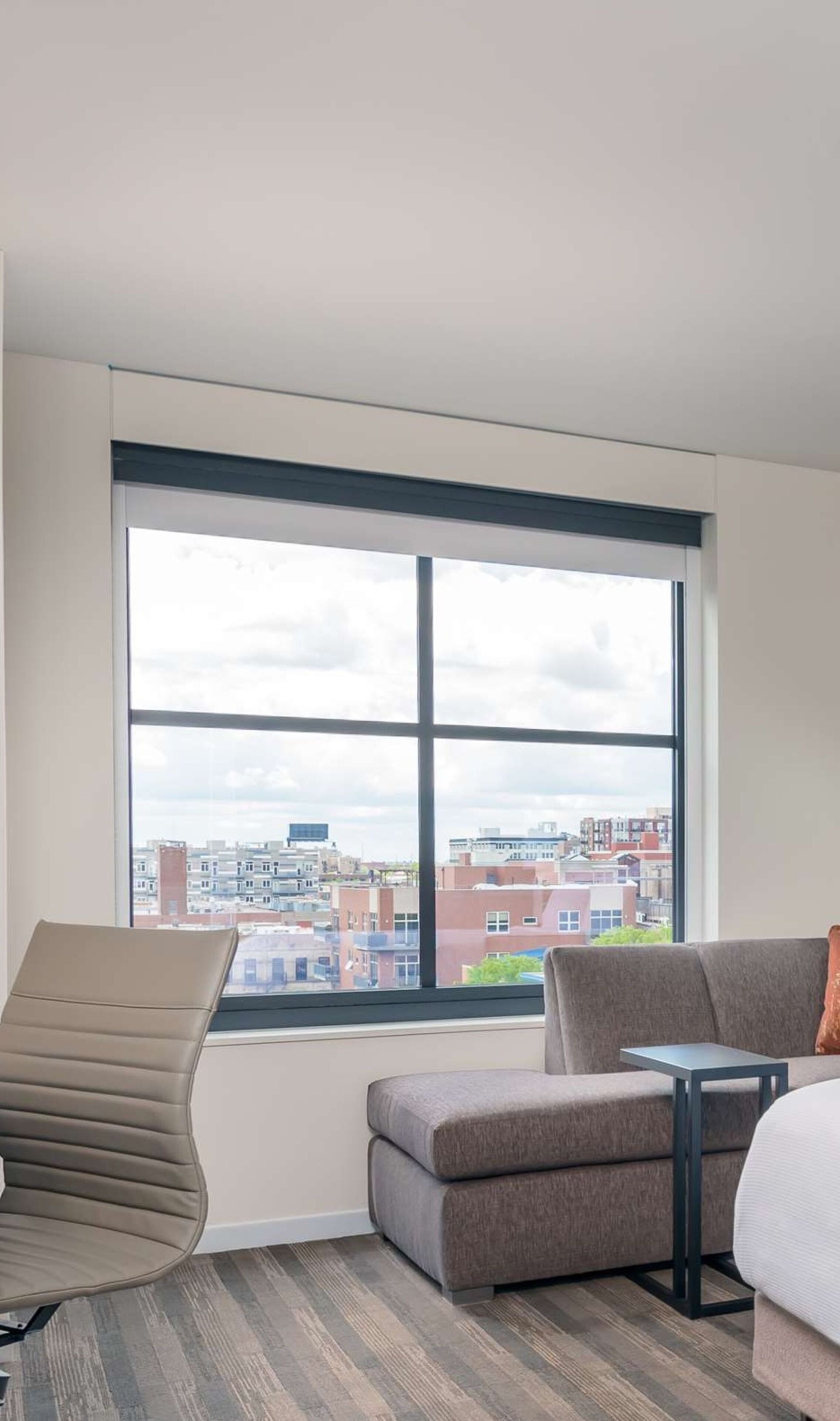 A modern hotel room with a large window, a sofa, a chair, and a small table.