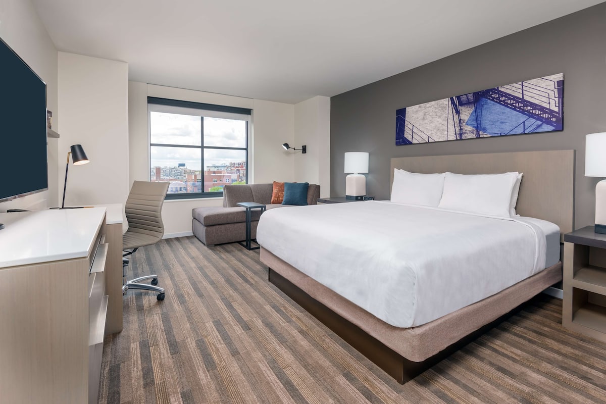 Une chambre d’hôtel moderne dotée d’un grand lit, d’un bureau, d’une chaise, d’un canapé et d’une fenêtre donnant sur la ville.