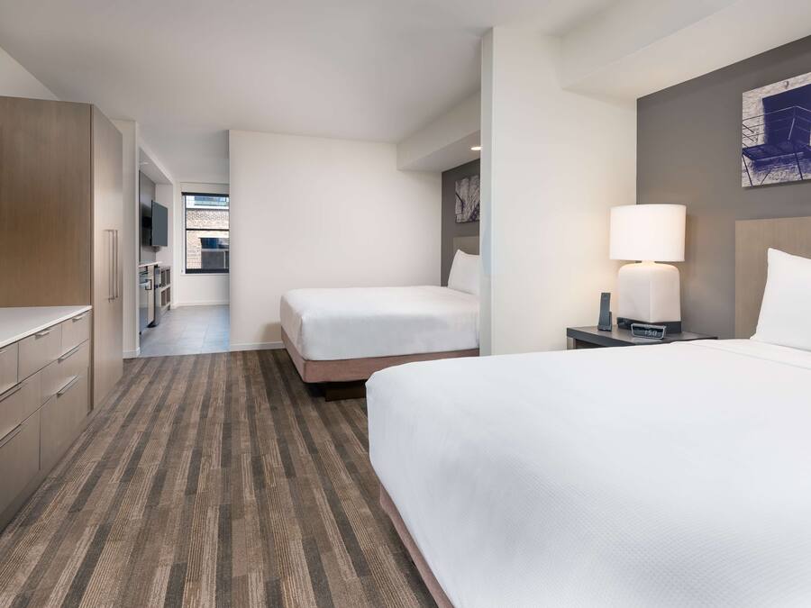 Habitación de hotel con una cama grande, una cama más pequeña, un escritorio con lámpara y televisión en la pared.