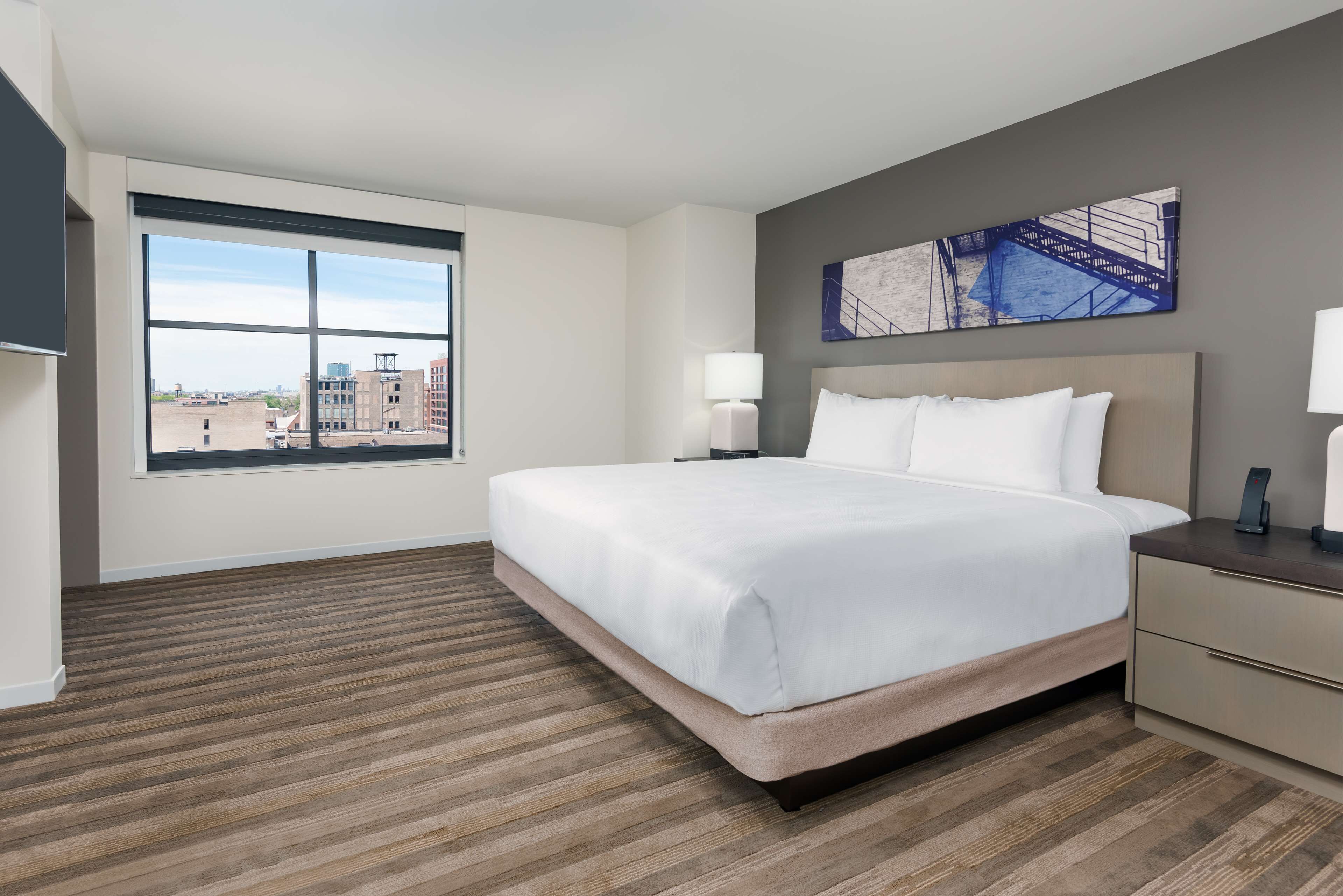 A modern hotel room with a large bed, a nightstand, a desk, and a window offering a city view.