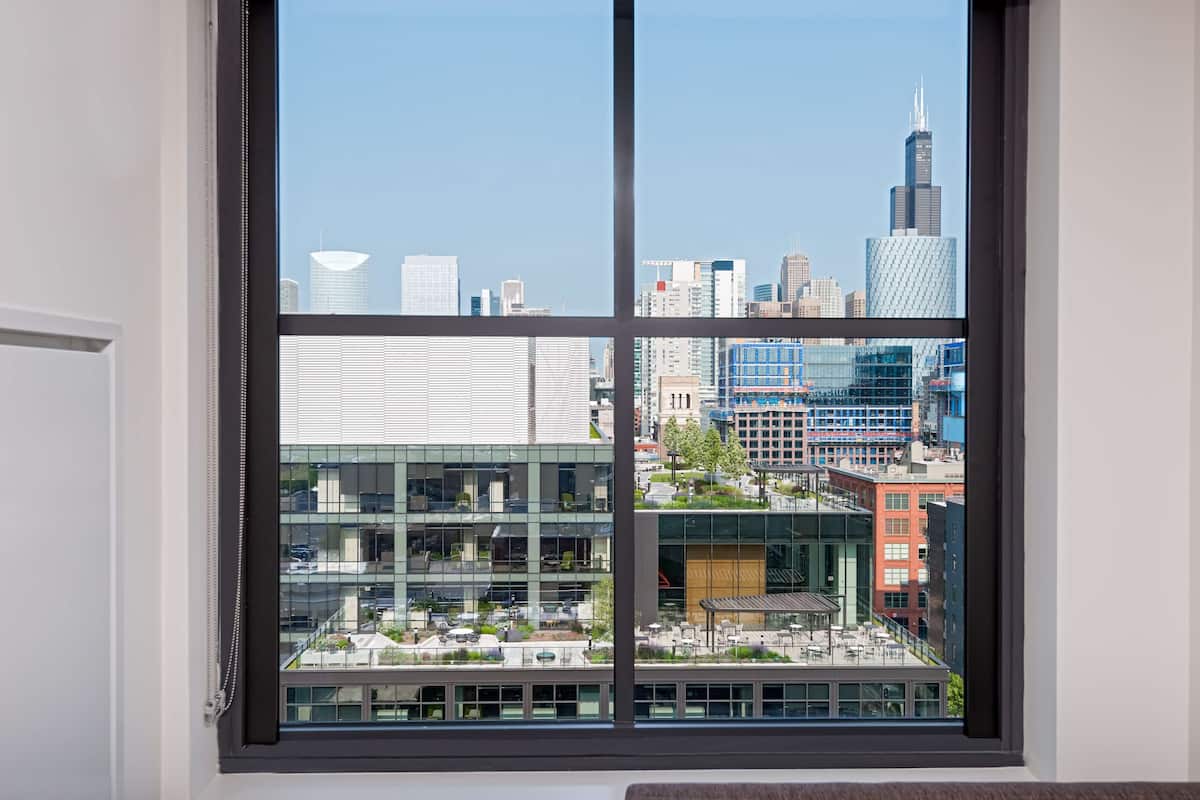 Une vue depuis une fenêtre, avec un paysage urbain en arrière-plan, comprenant des bâtiments modernes et un ciel dégagé.