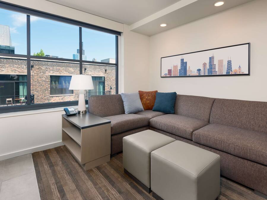 Una sala de estar moderna con un sofá modular, una mesita con una lámpara y vistas a un paisaje urbano a través de amplios ventanales.