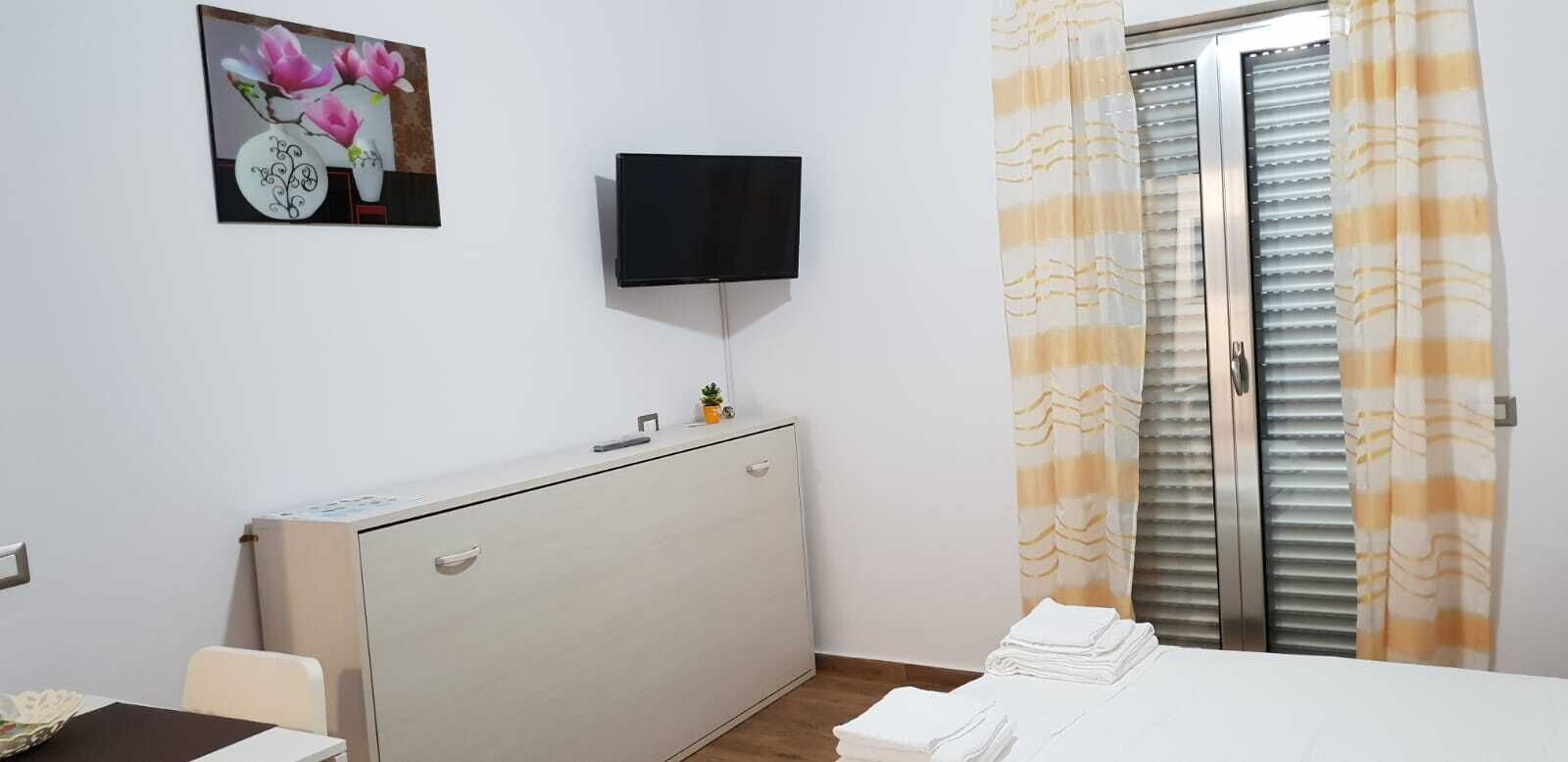 A bedroom with a bed, a desk, a TV mounted on the wall, and a window with curtains.