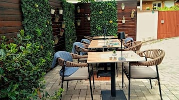 Un patio al aire libre con una mesa de madera y sillas de mimbre, rodeado de abundante vegetación y una valla de madera.