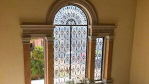 A spacious room with a large arched window featuring intricate ironwork, a potted plant, and a marble floor.
