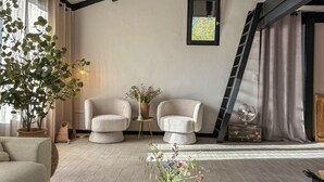 Ein gemütliches Zimmer mit zwei Sesseln, Holzboden, einem großen Fenster mit Vorhängen, einem kleinen Tisch mit Blumenvase und einer an der Wand stehenden Leiter.