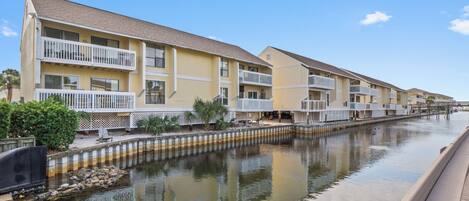 A row of yellow multi-story buildings with balconies, a canal in front, and a wooden boardwalk.