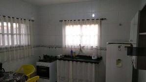 A compact kitchen with white tiled walls, a stove, a sink, and a refrigerator. There is a dining table with yellow chairs and lace curtains on the windows. - Itaipuaçu Residências Cajueiros II (Maricá)