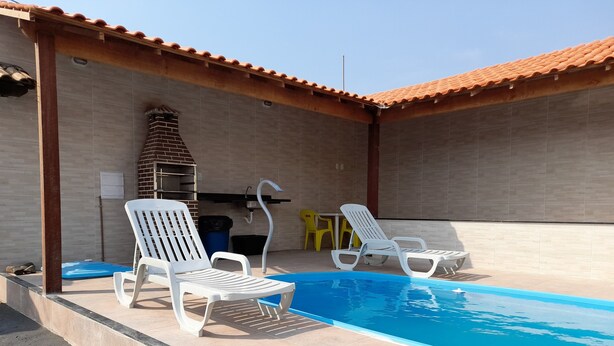 A swimming pool area with two white lounge chairs and a brick chimney. - Itaipuaçu Residências Cajueiros II (Maricá)