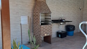 A brick-built outdoor grill with a chimney, potted plants, and a blue mat on a tiled patio. - Itaipuaçu Residências Cajueiros II (Maricá)