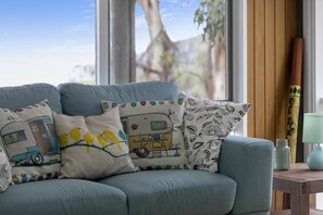 Un sofá azul con cojines estampados, una mesita de madera con una lámpara y una ventana con vistas a árboles.
