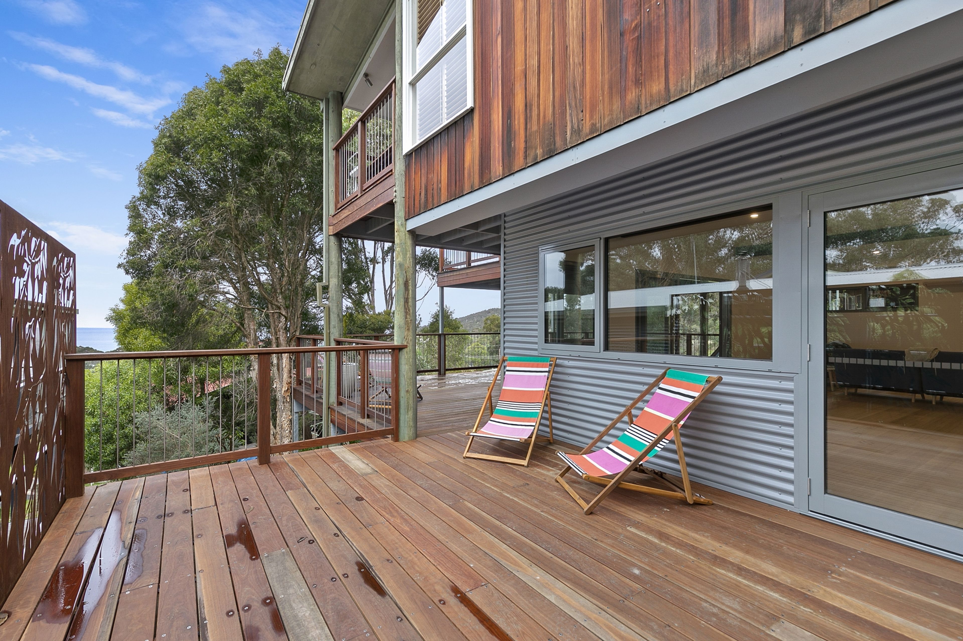 Une terrasse en bois avec deux chaises longues à rayures, une rambarde en métal et une vue sur un arbre et un bâtiment.