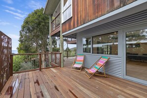 Une terrasse en bois avec deux chaises longues à rayures, une rambarde en métal et une vue sur un arbre et un bâtiment.