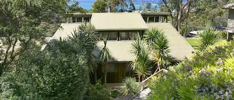 A two-story house with a flat roof, surrounded by dense vegetation and a view of the sea.
