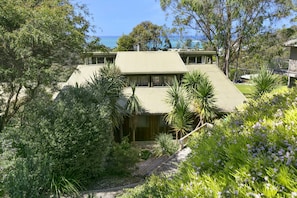 A two-story house with a flat roof, surrounded by dense vegetation and a view of the sea.