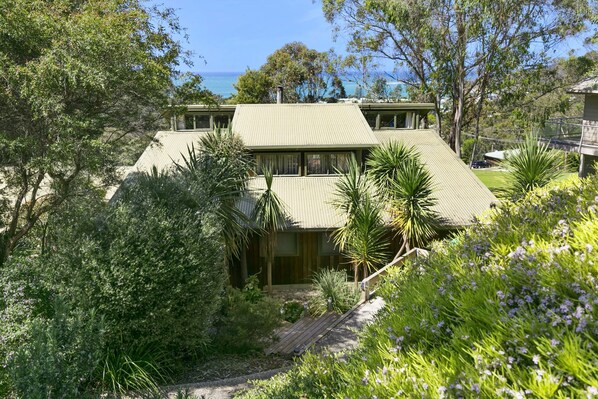 Ein zweigeschossiges Haus mit flachem Dach, umgeben von dichter Vegetation und Blick auf das Meer.