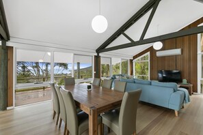 A modern living room with a wooden dining table, comfortable seating, a large TV, and a view of the outdoors.