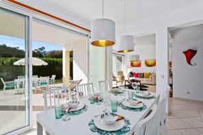 A dining area with a table set for four, a glass-enclosed patio, and a living room in the background. - Ocean Villas Villa Poetry (Albufeira)