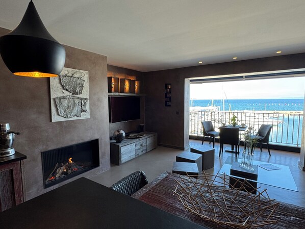 A modern living room with a fireplace, a flat-screen TV, and a balcony with a view of the sea.