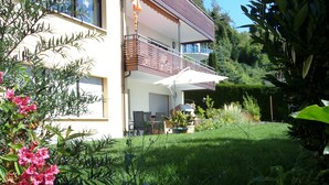 A modern two-story house with a balcony, a patio with outdoor furniture, and a well-maintained garden with various plants and flowers.