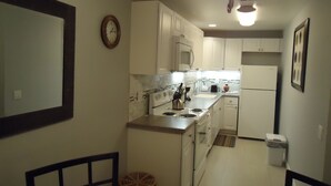 Une cuisine de taille réduite avec des meubles blancs, une cuisinière, un four à micro-ondes et un réfrigérateur. Il y a une horloge au mur et une chaise dans le coin.